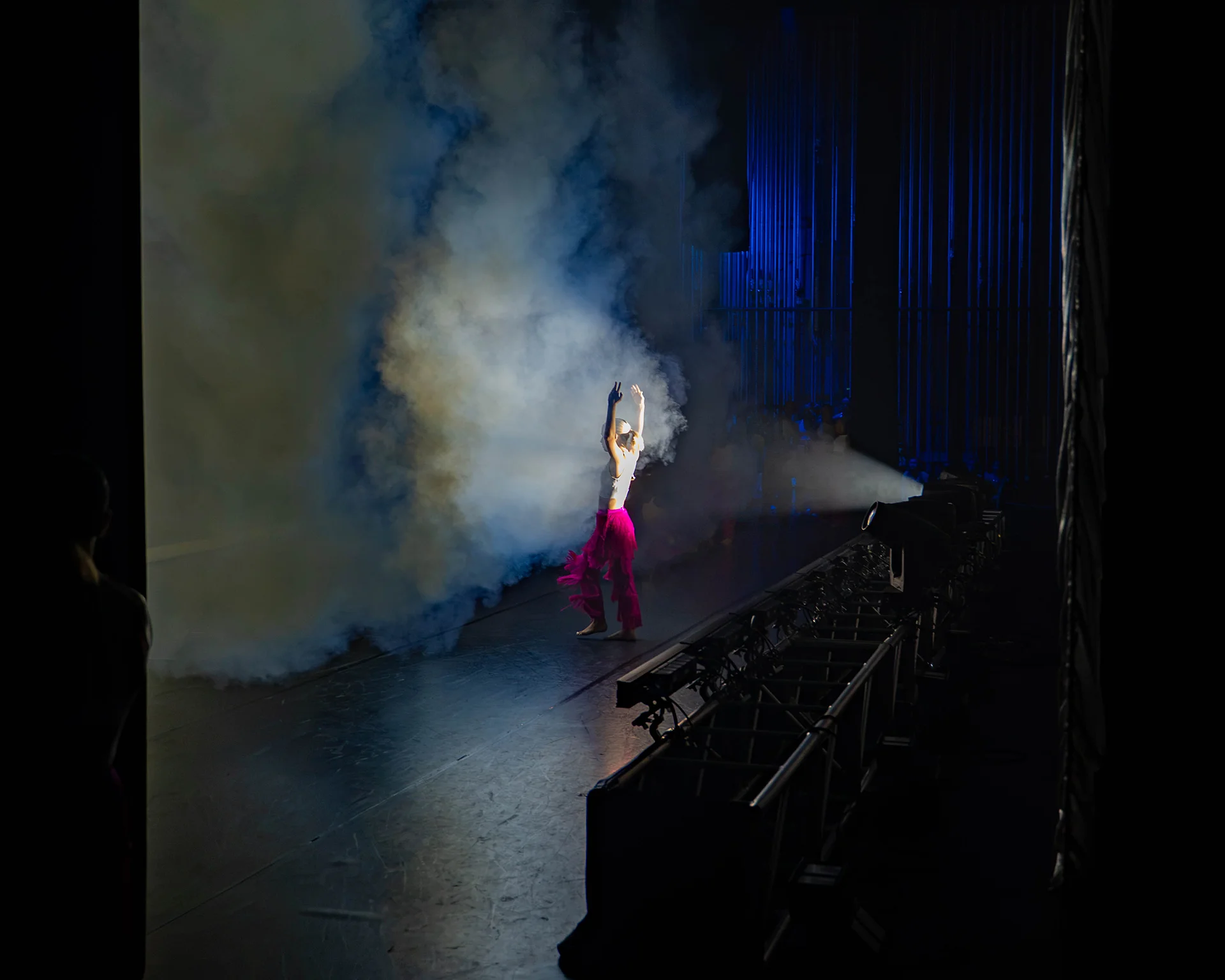 A performer raises their arms in a spotlight amid stage smoke and blue lighting.