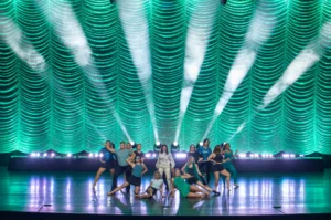 Dancers perform beneath white spotlights against a rippling teal curtain at a Bluewood Productions event.