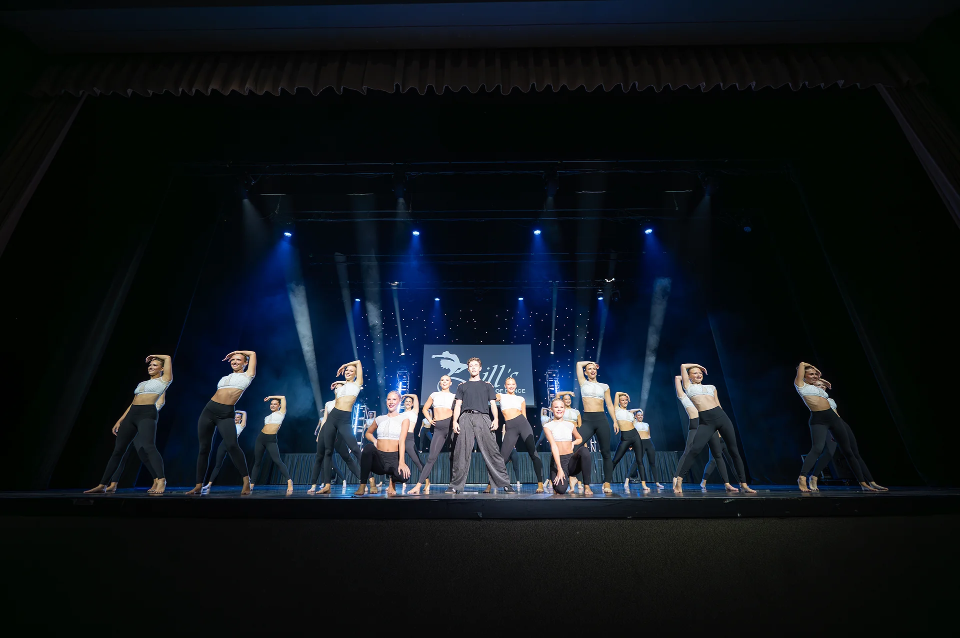 A dance troupe poses on a dark stage under blue spotlights.