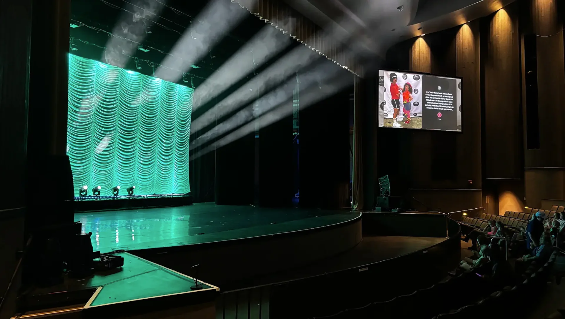 Bluewood Productions lights a stage with green drapery and white beams as attendees watch from the seats.