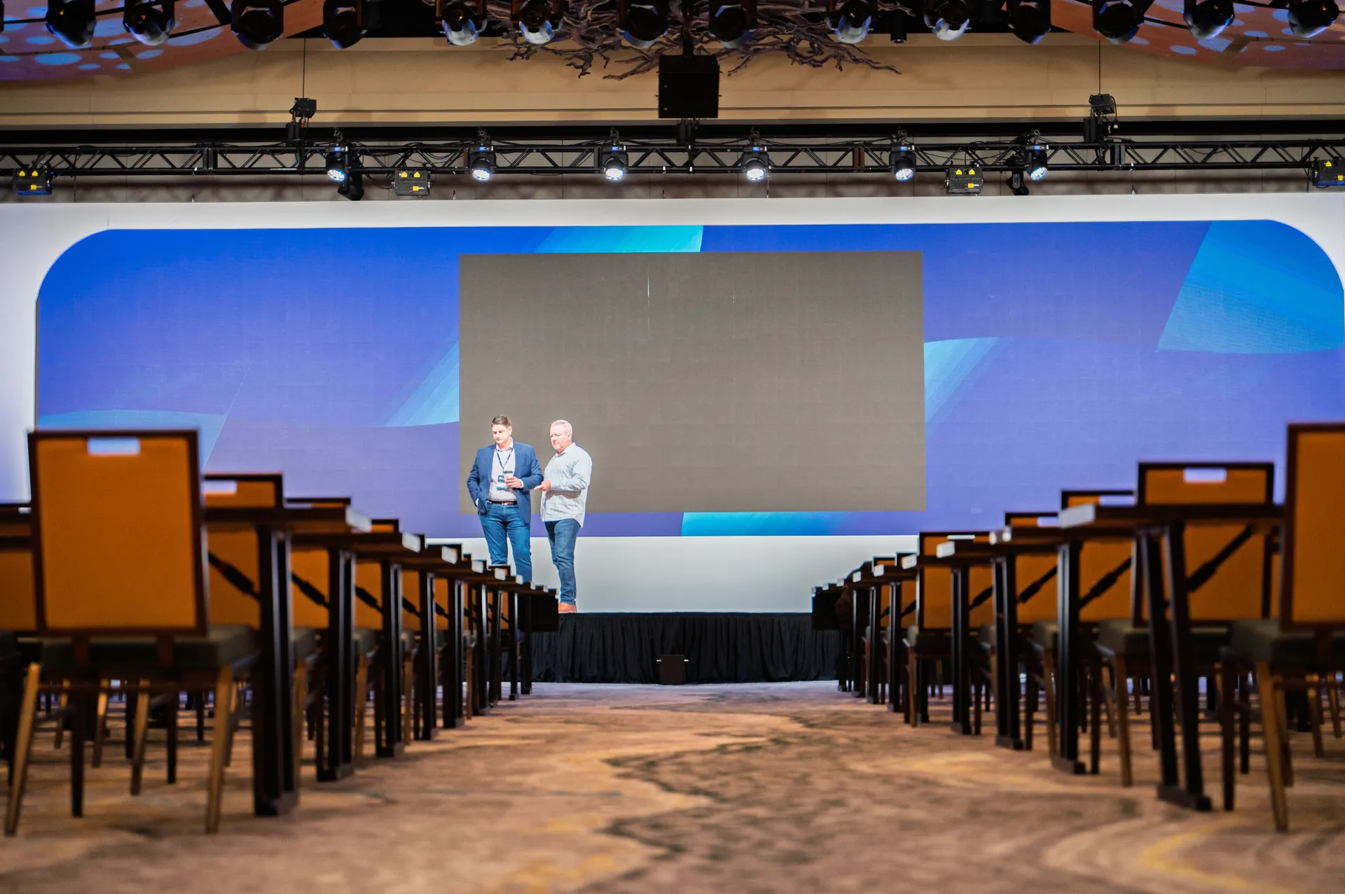 Two men stand on a stage, talking in front of a large screen in an empty conference room.